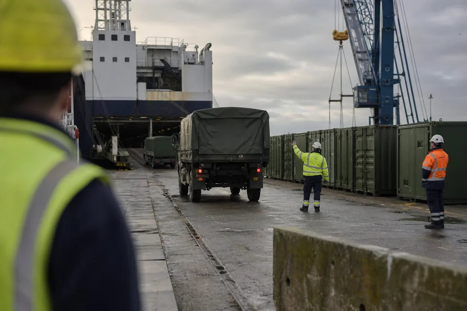 Military supply containers and vehicles being loaded onto a cargo ship at a port quay