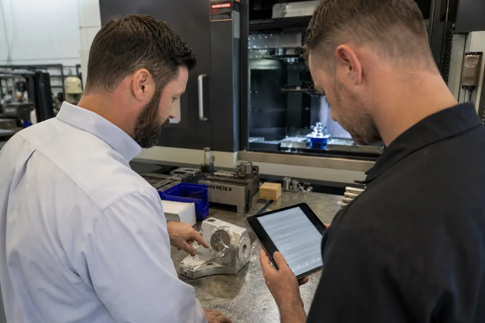 Two staff at a machining company comparing a tender list on a tablet next to a machined bracket